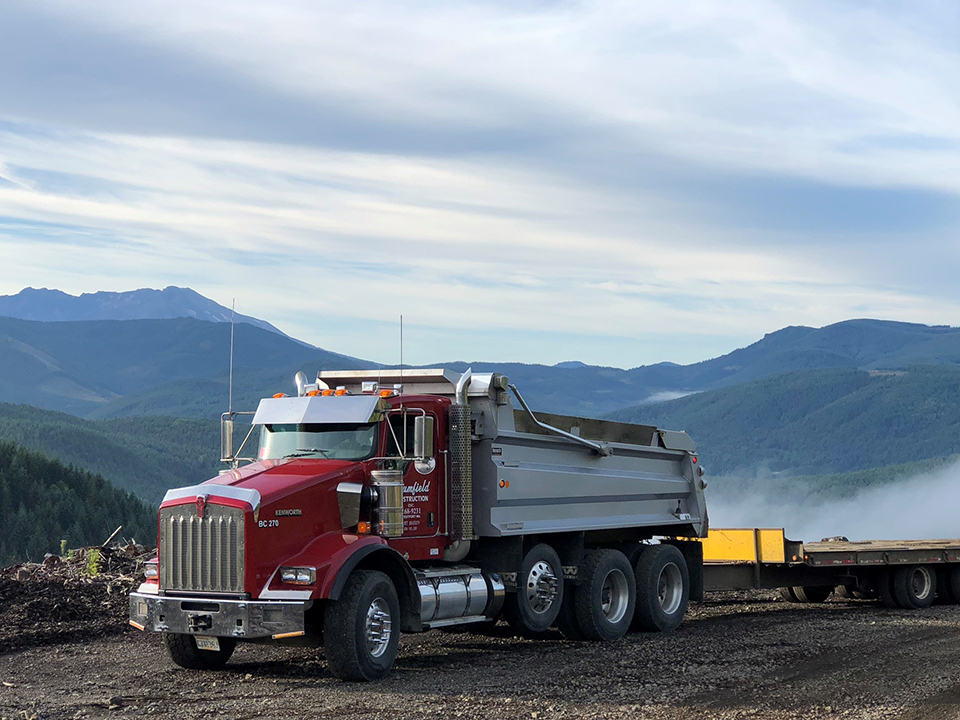 A red dump truck with the words "Brumfield Construction" on the side pulls a flatbed trailer along a dirt road in a mountainous landscape