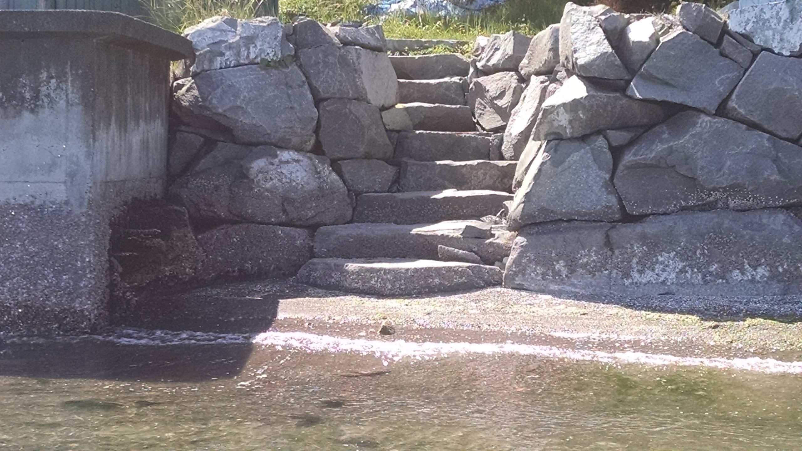 A stone bulkhead with uneven, moss-covered steps leading down to still water. The water is blue and reflects the clear sky